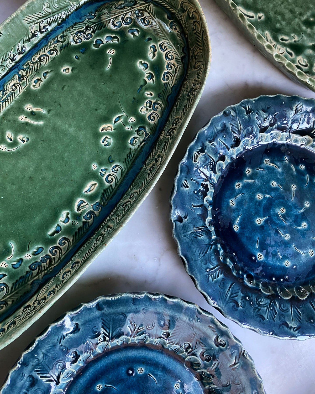 An overhead view of a green oval-shaped ceramic platter and two blue round-shaped ceramic platters, both with delicate impressions and surface patterns. TEST