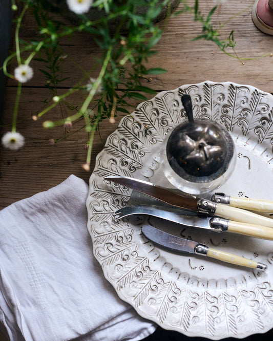 A close-up overhead view of a round handmade bohemian-style white ceramic platter with delicate impressions and surface patterns, complimented perfectly by vintage silverware, white linens and a rustic pine tabletop.