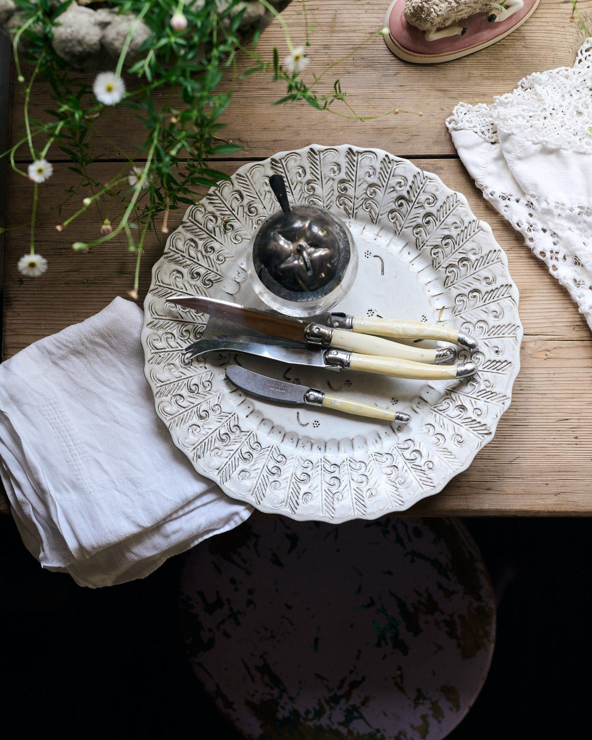 An overhead view of a round handmade bohemian-style white ceramic platter with delicate impressions and surface patterns, complimented perfectly by vintage silverware, white linens and a rustic pine tabletop. TEST