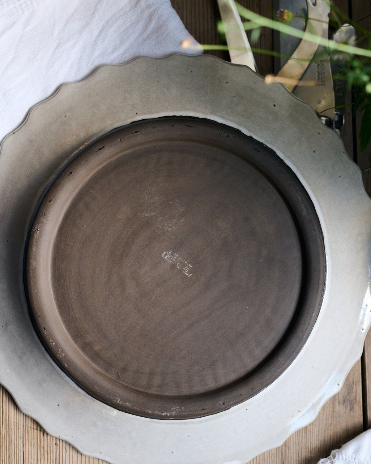 The underside of a round handmade bohemian-style ceramic platter. A white glazed rim surrounds an unglazed chocolate-coloured clay foot with a 'deVOL' logo in the centre. TEST