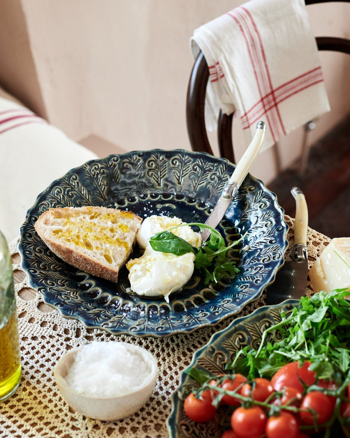 A round handmade bohemian-style blue ceramic platter, with cheese, tomatoes, homemade bread and leafy greens on a beautiful crochet lace tablecloth. TEST