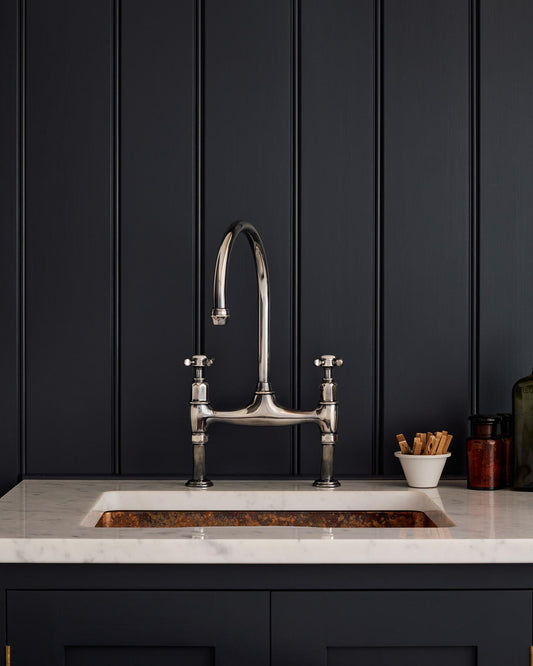 A straight-on view of a traditional-style tarnished silver mixer tap with crosshead handles in a classic utility kitchen with a Carrara marble worktop and deep blue painted cupboards and tongue and groove panelling.