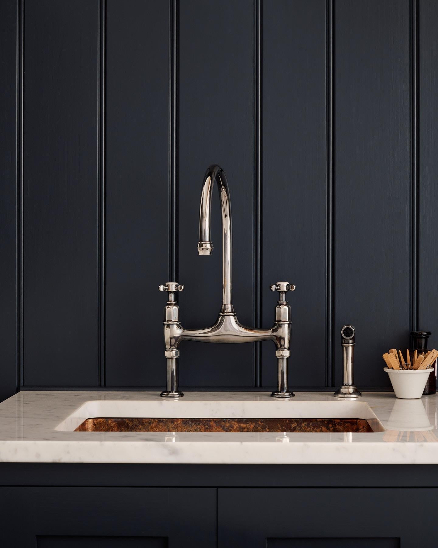 A straight-on view of a traditional-style tarnished silver mixer tap with crosshead handles and an independent rinser tap in a classic utility kitchen with a Carrara marble worktop and deep blue painted cupboards and tongue and groove panelling.