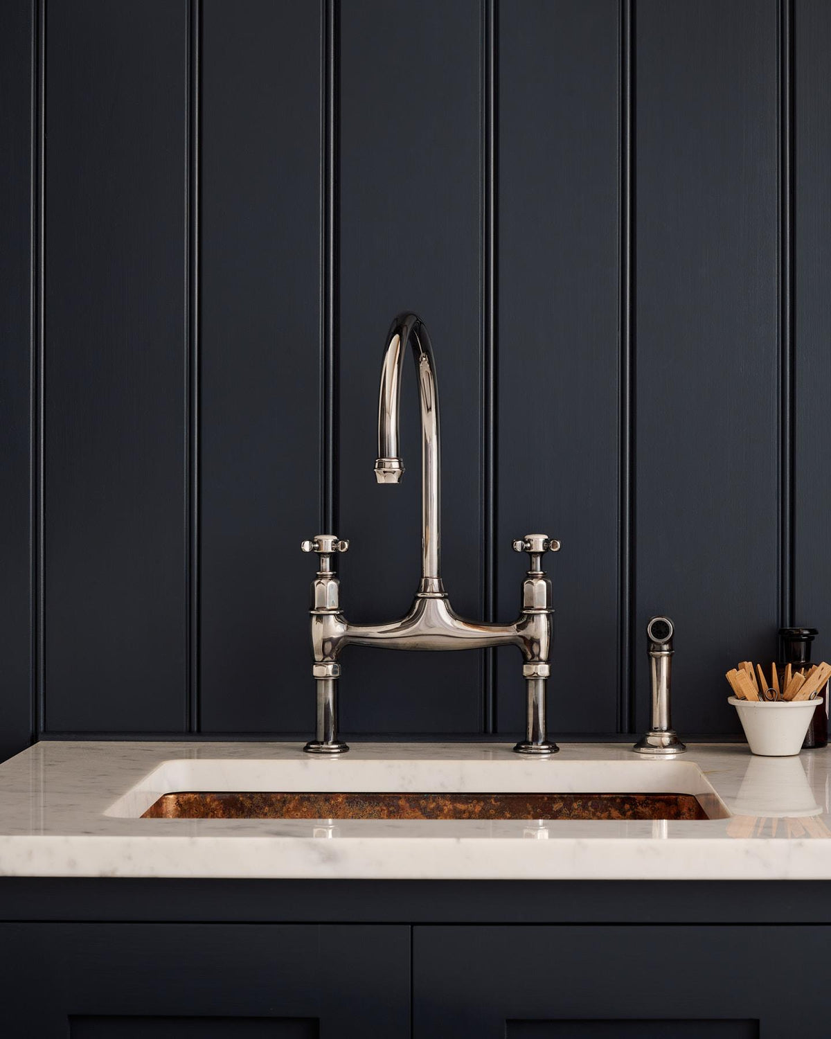 A straight-on view of a traditional-style tarnished silver mixer tap with crosshead handles and an independent rinser tap in a classic utility kitchen with a Carrara marble worktop and deep blue painted cupboards and tongue and groove panelling. TEST