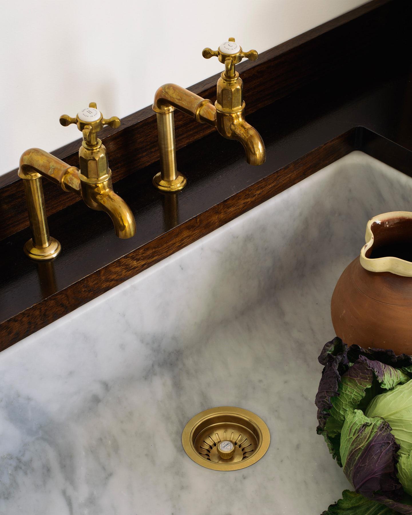 A pair of traditional-style aged brass bibcock taps with crosshead handles seen from overhead. They are fitted to a dark hardwood worktop over an elegant Carrara marble sink.