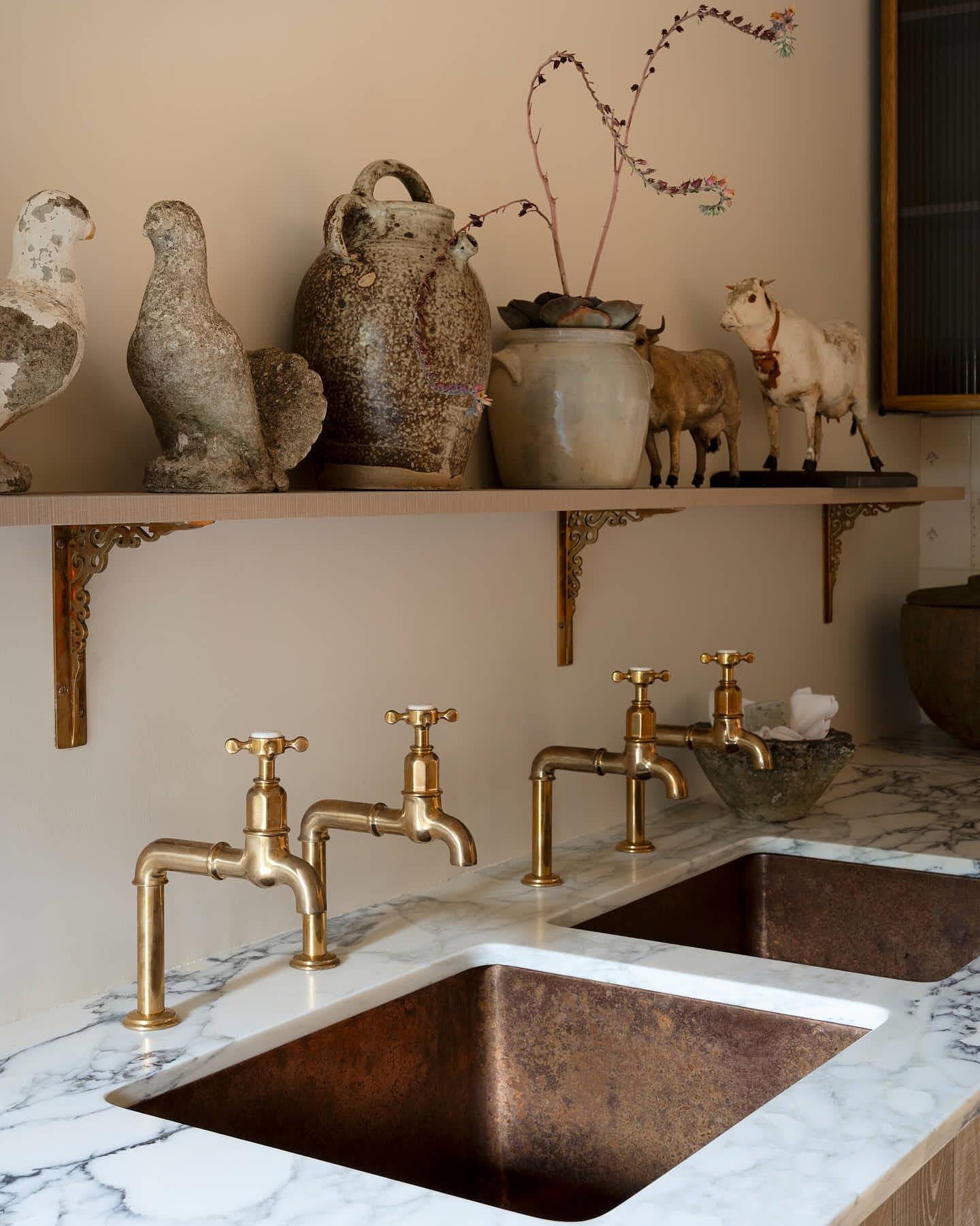 A close-up view of a pair of traditional-style aged brass bibcock taps with crosshead handles. An 'H' can be seen in the centre of the handle with the words 'deVOL' and 'Made in England' around the perimeter.