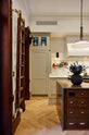 A grand and spacious classic English kitchen features a half vintage and half deVOL kitchen island with a copper sink and worktop, a cooker run with an Arabescato marble worktop, and a full-height cupboard. Beautifully decorated with artisan glass and ceramics, a sophisticated floral arrangement, an aged brass hanging rail, a bespoke ladder, and milk glass pendant lights, it creates a social space with a touch of luxury.