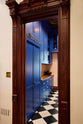 Beyond an ornate wooden doorway lies a walk-in galley pantry that boasts full-height cupboards, wall cupboards, and a drawer base cabinet topped with solid oak. This pantry is beautifully decorated with artisan ceramics, a bowl of artichokes, and dried ingredients stored in clear containers, all set against checkered marble tiles. The thoughtful design creates a social space that exudes a touch of luxury, making it perfect for both cooking and entertaining.