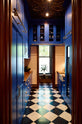 Beyond an ornate wooden doorway is a walk-in galley pantry featuring full-height cupboards, wall cupboards, and a small sink run with an Arabescato marble basin. Topped with solid oak, the drawer base cabinet complements the decor of artisan ceramics, a bowl of artichokes, and clear containers of dried ingredients, all set against checkered marble tiles. This thoughtful design creates a luxurious social space, perfect for cooking and entertaining.