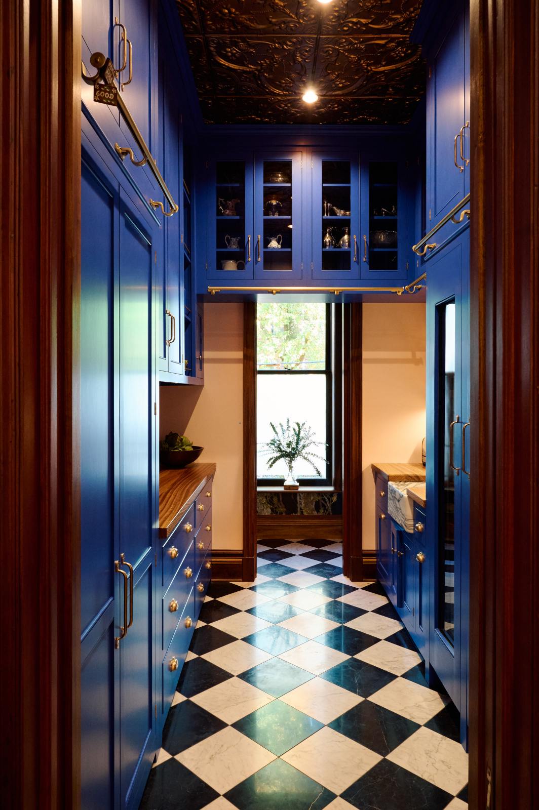 Beyond an ornate wooden doorway is a walk-in galley pantry featuring full-height cupboards, wall cupboards, and a small sink run with an Arabescato marble basin. Topped with solid oak, the drawer base cabinet complements the decor of artisan ceramics, a bowl of artichokes, and clear containers of dried ingredients, all set against checkered marble tiles. This thoughtful design creates a luxurious social space, perfect for cooking and entertaining.
