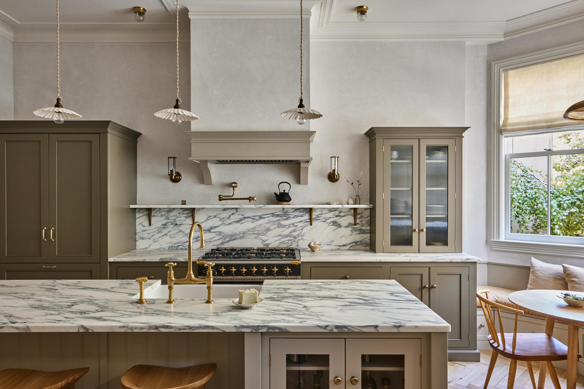 An elegant mushroom-brown, classic English kitchen features a cooker run with an Arabescato marble worktop, splashback, and shelf; a reeded countertop cupboard; a full-height pantry cupboard; and a large kitchen island with a fluted Belfast sink and an aged brass Ionian tap. Beautifully decorated with artisan ceramics, narrow pleated porcelain lights, deVOL Bum Stools, and a cast-iron teapot, it adds a sophisticated touch to the elegant kitchen.