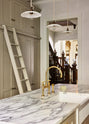An angled look at an elegant mushroom-brown, classic English kitchen features a large kitchen island with a fluted Belfast sink, an aged brass Ionian tap, Arabescato marble worktop, and a full-height pantry cupboard with a ladder. Beautifully decorated with artisan ceramic, narrow pleated porcelain lights, and a bar of hand-made soap, it adds a sophisticated touch to the elegant kitchen.