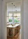 An angled look at an elegant mushroom-brown, classic English kitchen features a large kitchen island with a fluted Belfast sink, an aged brass Ionian tap, and an Arabescato marble worktop. Beautifully decorated with artisan ceramics, narrow pleated porcelain lights, and wooden deVOL bum stools, it adds a sophisticated touch to the elegant kitchen.