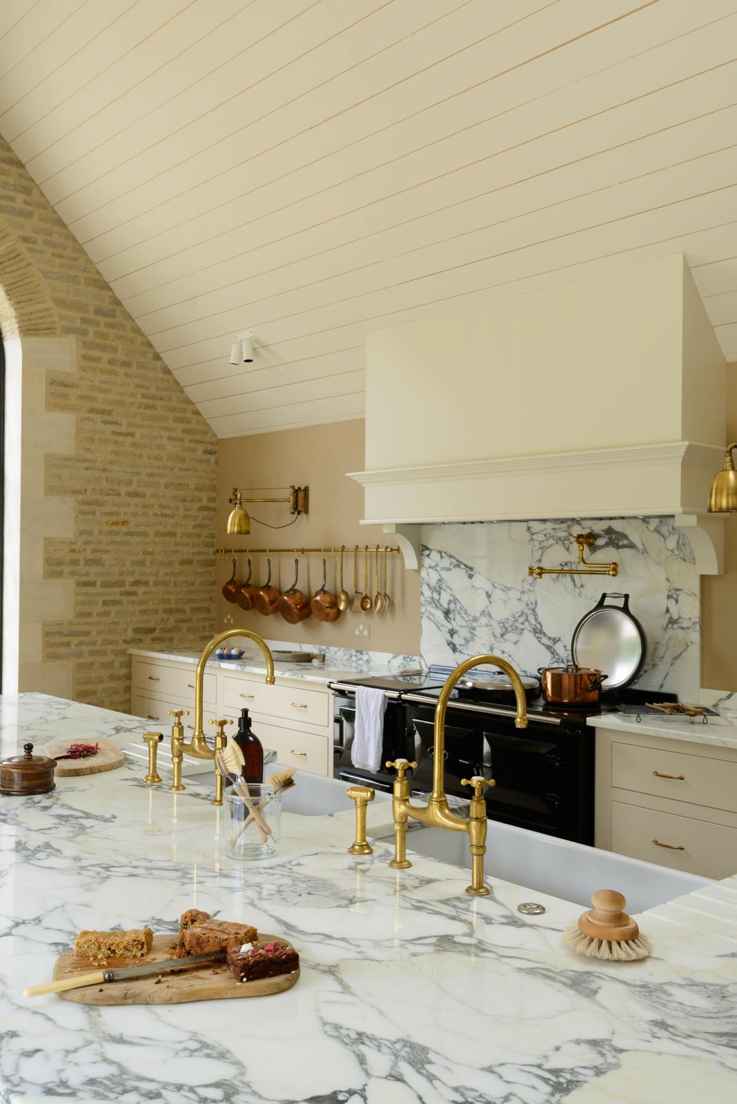 An oblique view of a grand mushroom-coloured classic English kitchen features a cooker run with a suspended chimney breast, a large kitchen island, two sets of sinks, aged brass Ionian taps, and an Arabescato marble worktop. Beautifully decorated with aged brass hanging rails, simple ceramics, brass folded wall lamps, and a large collection of kitchen utensils, this space creates a humble family atmosphere despite its luxurious design.