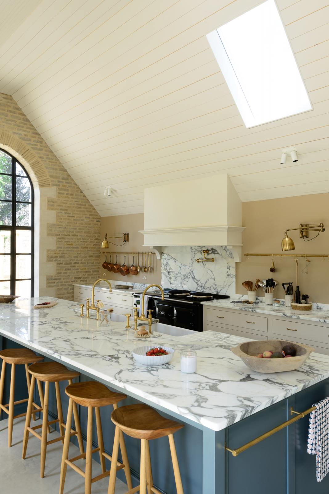 An oblique view of a grand mushroom-coloured classic English kitchen features a cooker run with a suspended chimney breast, a large kitchen island, two sets of sinks, aged brass Ionian taps, and an Arabescato marble worktop. Beautifully decorated with bum stools, aged brass hanging rails, simple ceramics, brass folded wall lamps, and a large collection of kitchen utensils, this space creates a humble family atmosphere despite its luxurious design.