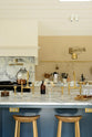 A grand mushroom-coloured classic English kitchen features a cooker run with a suspended chimney breast, a large kitchen island, two sets of sinks, aged brass Ionian taps, and an Arabescato marble worktop. Beautifully decorated with a aged brass hanging rail, bum stools, simple ceramics, a brass folded wall lamp, and a large collection of kitchen utensils, this space creates a humble family atmosphere despite its luxurious design.
