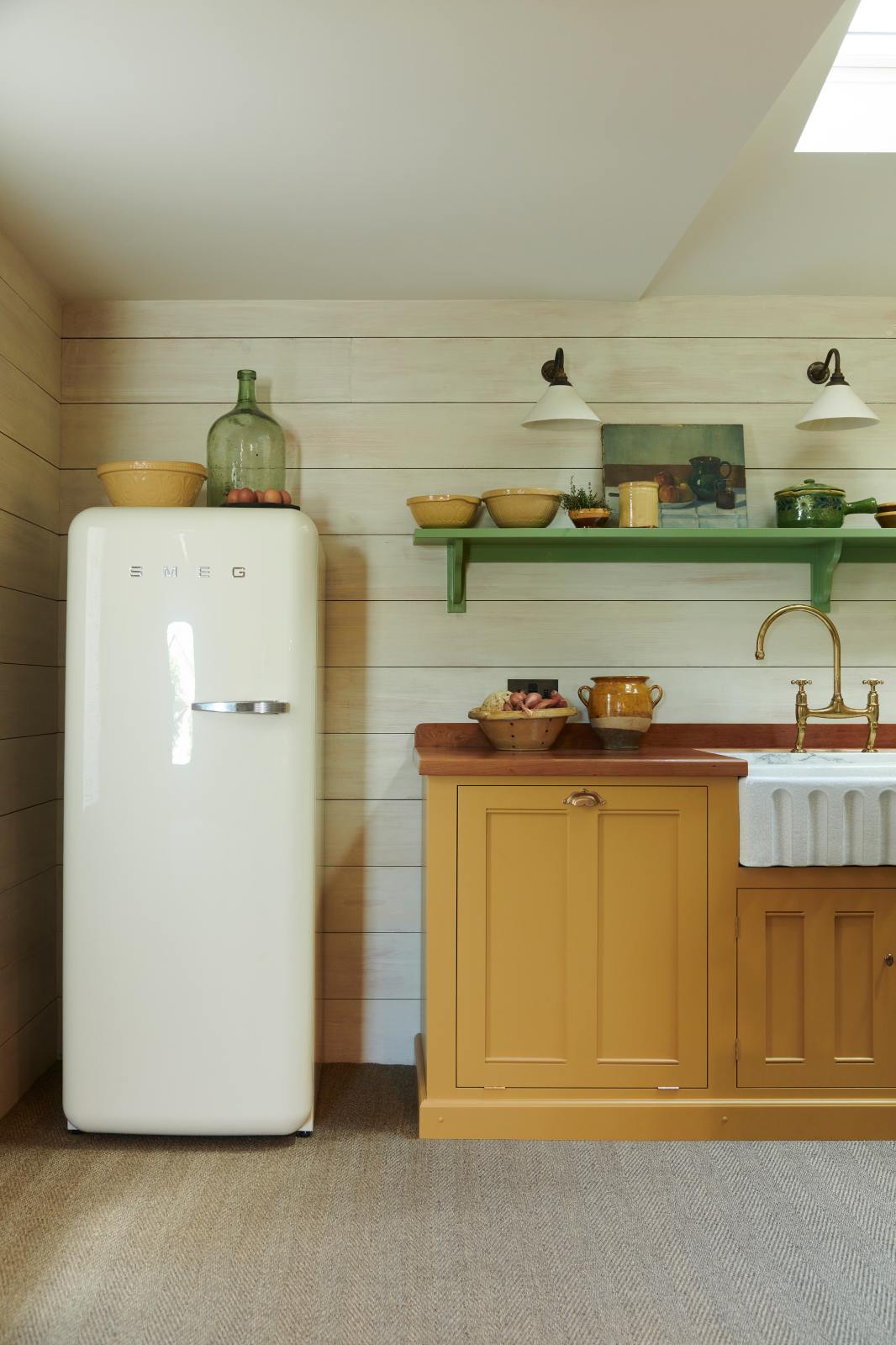 A quiet corner of a quaint and glamorous cottage kitchen, in shades of green and scullery yellow, includes a small sink run with an oak countertop, an aged brass Ionian tap, and a Smeg fridge. Lovingly decorated with vintage furniture, ceramics, luscious vegetables, and three deVOL mini creamware wall lights, the space exudes personality and charm while respecting the cottage's original textures.