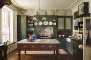 An eccentric yet grand Bakehouse Green Classic English Kitchen features an L-shaped sink and cooker run with black granite worktop, a glazed wall cupboard, an overmantle with cream subway splashback tiles, a full-height cabinet, and an Heirloom dairy table. Pridefully displaying taxidermy, artisan ceramics, and collectable antiques, it embraces a unique museum-like social kitchen space.