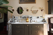 A glamorous Bakehouse green bathroom with a bespoke sink cabinet with a marble worktop, shelf and splashback. Beautifully detailed cream wallpaper, a simple mirror, an elegant painting, and a touch of greenery from a houseplant come together to create a gorgeously chic space.