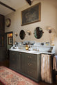 An oblique view of a glamorous Bakehouse green bathroom with a bespoke sink cabinet with a marble worktop, shelf and splashback. Beautifully detailed cream wallpaper, a simple mirror, an elegant painting, and a touch of greenery from a houseplant come together to create a gorgeously chic space.