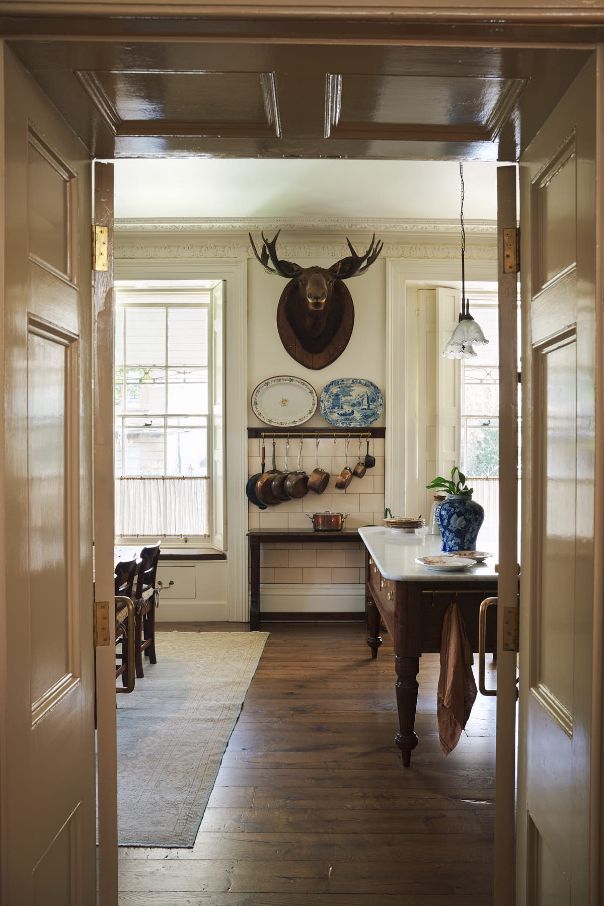 Looking through an open door way is an eccentric yet grand Bakehouse Green Classic English Kitchen features an oiled oak shelf with brass hanging rail, and an Heirloom dairy table. Pridefully displaying a taxidermy moose, antique furniture, and artisan ceramics, it embraces a unique museum-like social kitchen space.