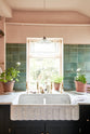 A closer look at the bright peach-coloured kitchen reveals a Carrara marble worktop with a double fluted sink and twin aged brass Mayan taps. Vintage teal lace-market splashback tiles add character to the space. The space is styled with a radio, potted plants scattered across the counter, and a large plant on the floor.