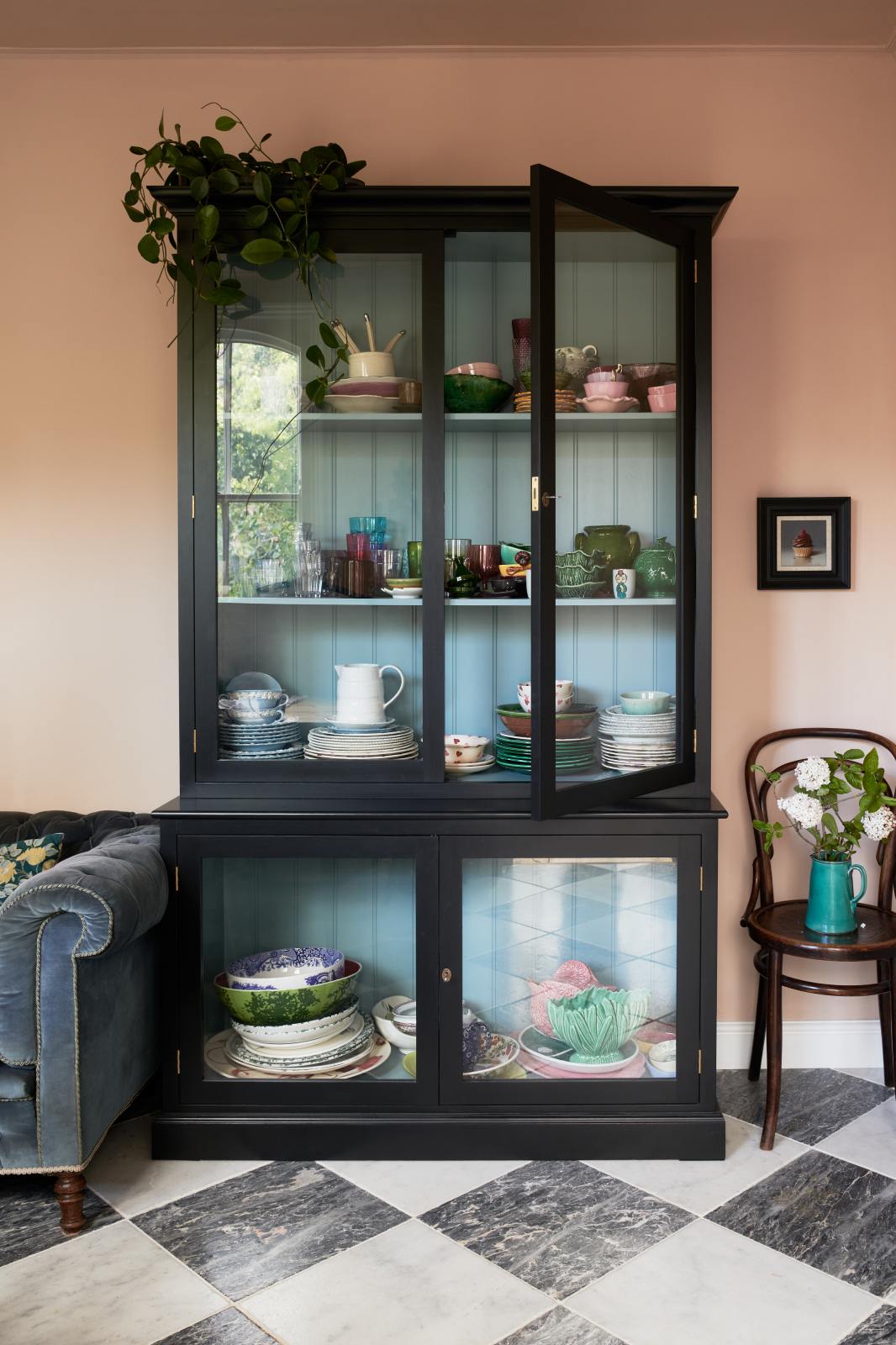 A closer look at the painter’s black curiosity cupboard reveals a Trinity Blue interior that beautifully highlights a collection of quirky ceramic plates, bowls, and mugs. The bright peach-coloured room features a chequerboard marble floor and an eclectic collection of vintage furniture, including a velvet blue sofa, a small cupcake painting, blossoming branches in a teal jug, and a wooden chair.