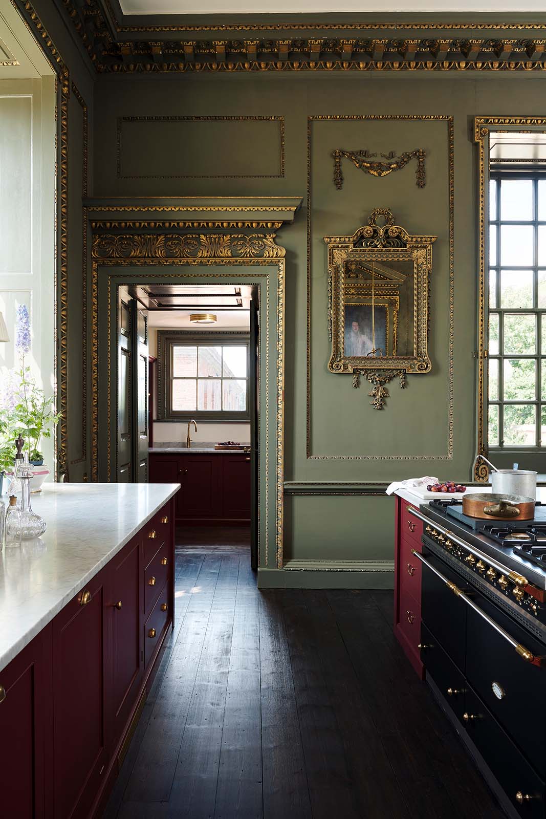 A grand, refectory red classic English kitchen featuring a sink run with Carrara marble worktop, a kitchen island, and a small scullery. Place upon a tall, magnificent open space with deep olive green walls with ornate gold gilding, and beautifully decorated with a Gaselier light, artisan ceramics, and vintage furniture, creating a splendidly royal and practical use of space.