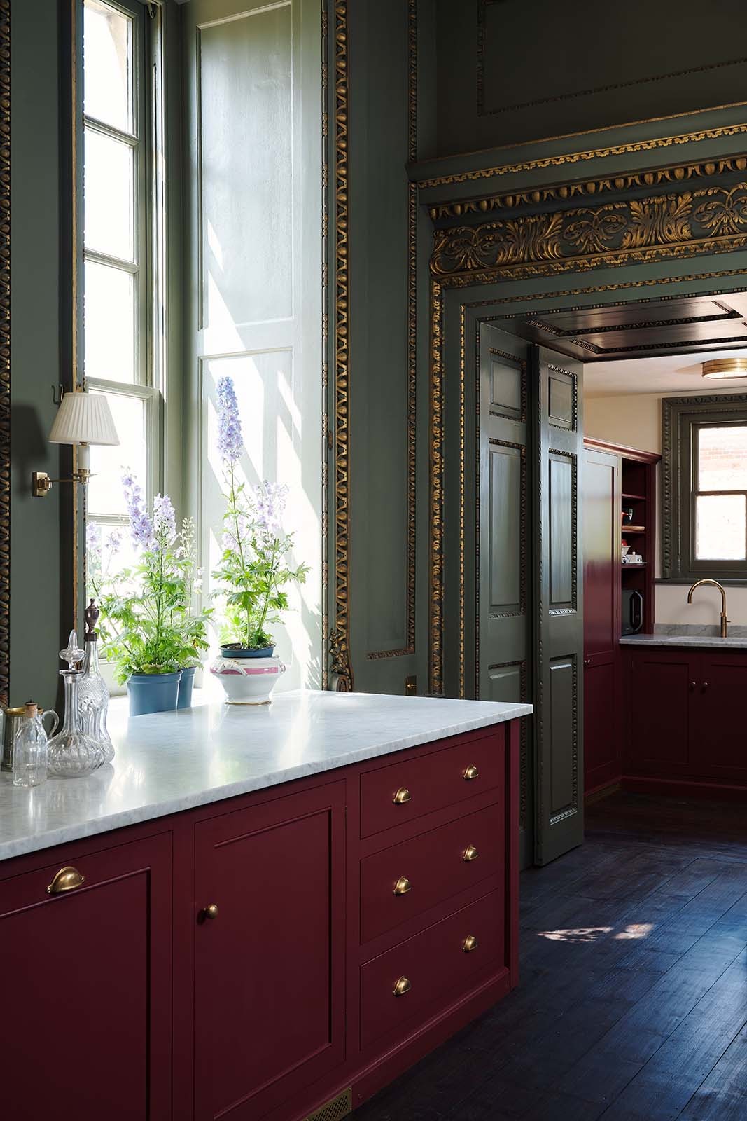 A quiet corner of a grand, refectory red classic English kitchen featuring a sink run with Carrara marble worktop, a kitchen island, and a tall pantry cupboard. Place upon a tall, magnificent open space with deep olive green walls with ornate gold gilding, and beautifully decorated with a Gaselier light, artisan ceramics, and vintage furniture, creating a splendidly royal and practical use of space.