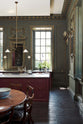 A quiet corner of a grand, refectory red classic English kitchen featuring a sink run  with Carrara marble worktop, a kitchen island, and a tall pantry cupboard. Place upon a tall, magnificent open space with deep olive green walls with ornate gold gilding, and beautifully decorated with a Gaselier light, artisan ceramics, and vintage furniture, creating a splendidly royal and practical use of space.