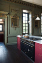 An oblique view of a grand, refectory red classic English kitchen featuring a kitchen island with Carrara marble worktop. Place upon a tall, magnificent open space with deep olive green walls with ornate gold gilding, and beautifully decorated with a Gaselier light, artisan ceramics, and vintage furniture, creating a splendidly royal and practical use of space.