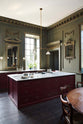 An oblique view of a grand, refectory red classic English kitchen featuring a sink run and cooker with Carrara marble worktop, and a kitchen island. Place upon a tall, magnificent open space with deep olive green walls with ornate gold gilding, and beautifully decorated with a Gaselier light, artisan ceramics, and vintage furniture, creating a splendidly royal and practical use of space.