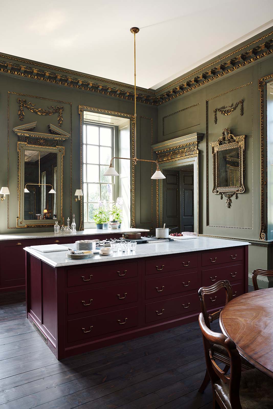 An oblique view of a grand, refectory red classic English kitchen featuring a sink run and cooker with Carrara marble worktop, and a kitchen island. Place upon a tall, magnificent open space with deep olive green walls with ornate gold gilding, and beautifully decorated with a Gaselier light, artisan ceramics, and vintage furniture, creating a splendidly royal and practical use of space.