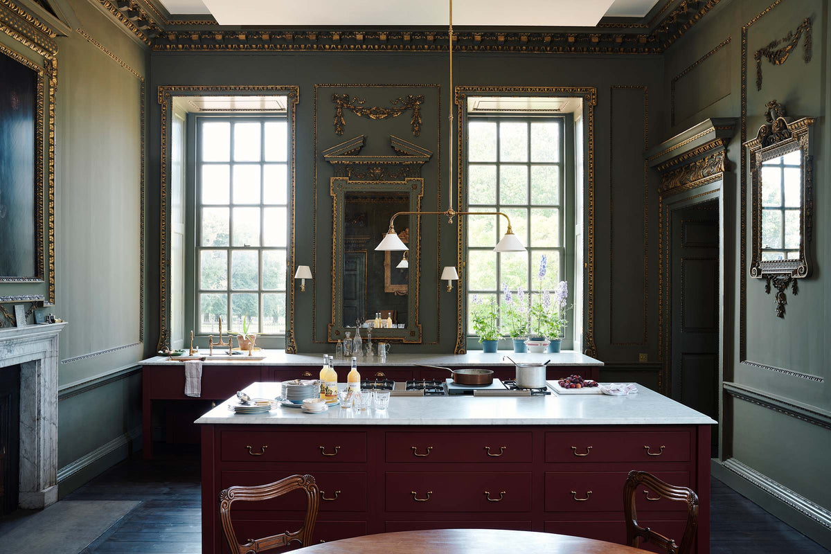 A grand, refectory red classic English kitchen featuring a sink run and cooker with Carrara marble worktop, and a kitchen island. Place upon a tall, magnificent open space with deep olive green walls with ornate gold gilding, and beautifully decorated with a Gaselier light, artisan ceramics, and vintage furniture, creating a splendidly royal and practical use of space.