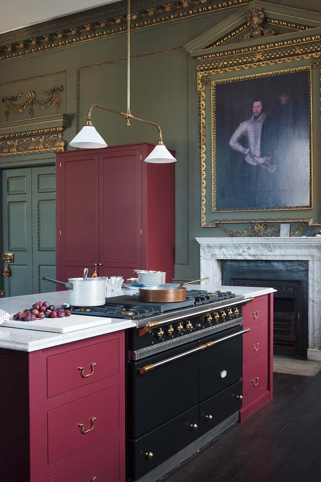 An oblique view of a grand, refectory red classic English kitchen featuring a kitchen island with Carrara marble worktop, and a tall pantry cupboard. Place upon a tall, magnificent open space with deep olive green walls with ornate gold gilding, and beautifully decorated with a Gaselier light, artisan ceramics, and vintage furniture, creating a splendidly royal and practical use of space.