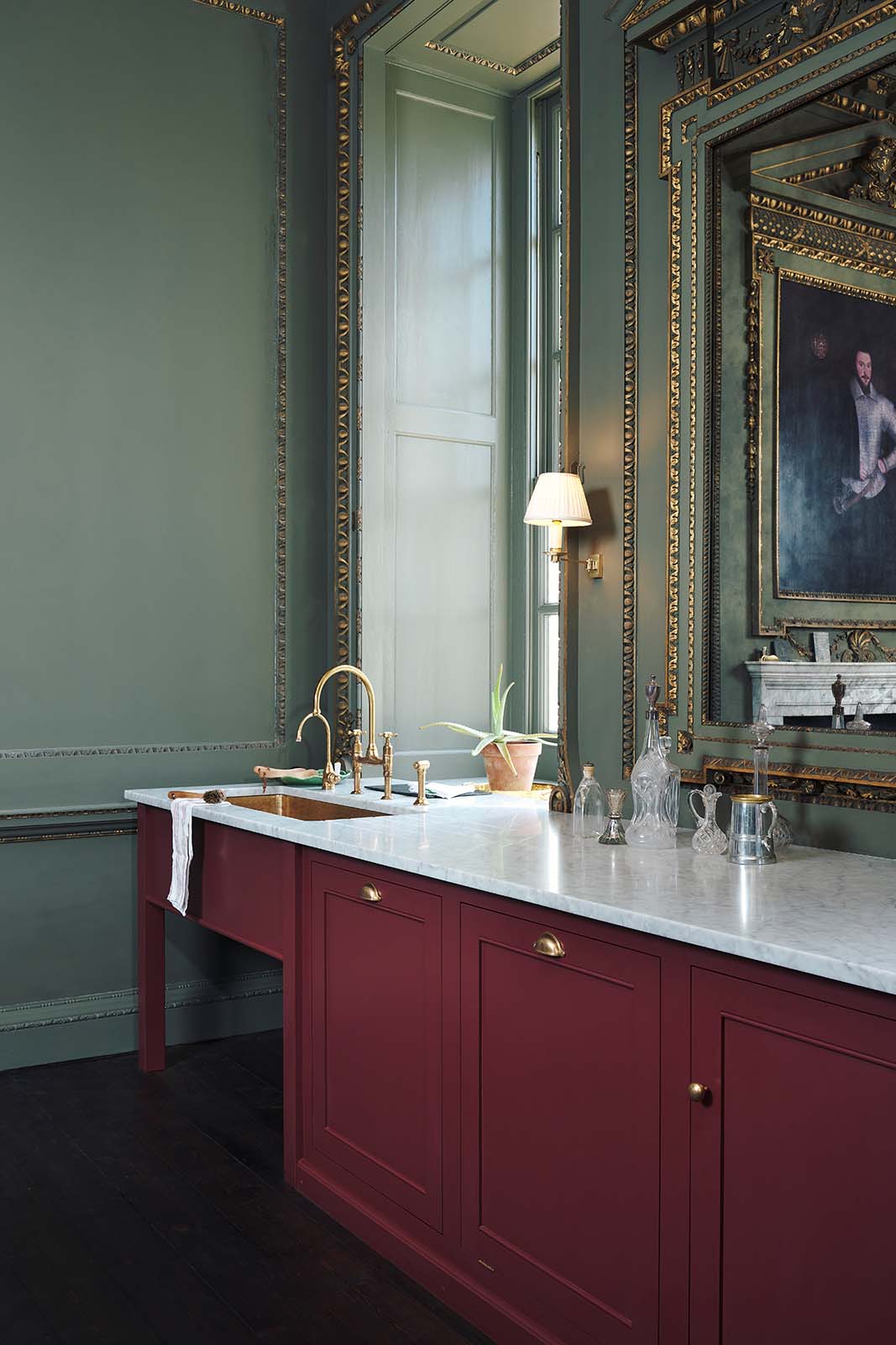 An oblique view of a quiet corner of a grand, refectory red classic English kitchen featuring a sink run with Carrara marble worktop, an embedded brass sink, and an aged brass Ionian tap. Place upon a tall, magnificent open space with deep olive green walls with ornate gold gilding, and beautifully decorated with a Gaselier light, artisan ceramics, and vintage furniture, creating a splendidly royal and practical use of space.