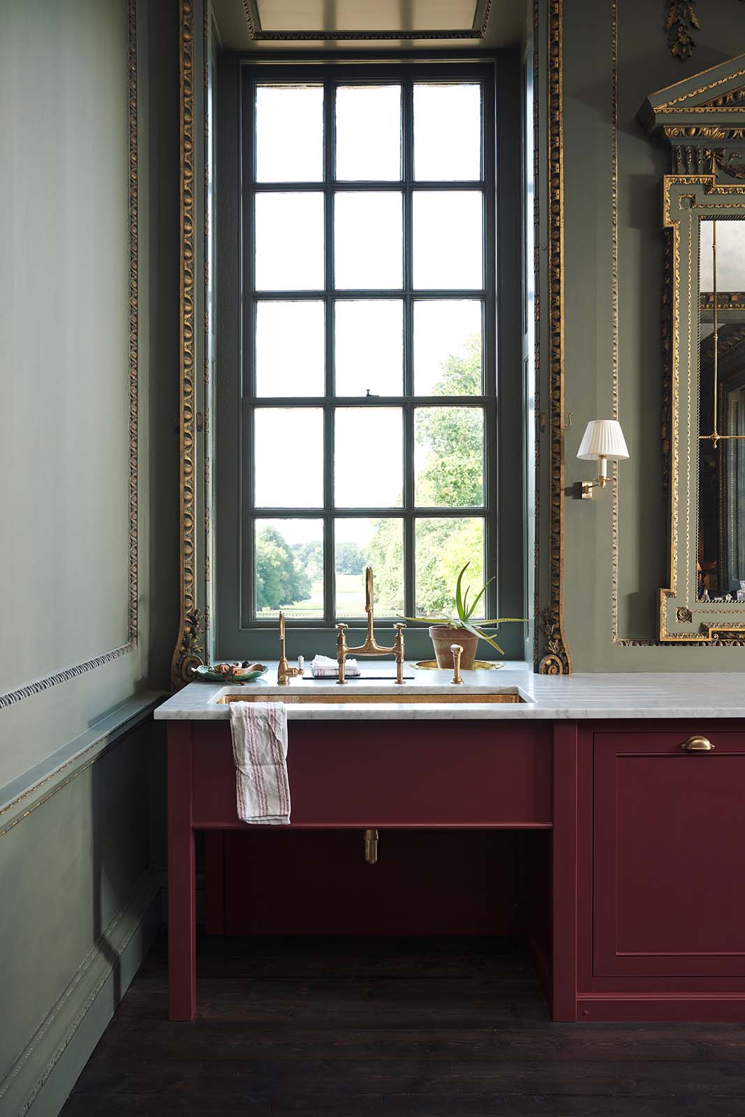 A quiet corner of a grand, refectory red classic English kitchen featuring a sink run with Carrara marble worktop, an embedded brass sink, and an aged brass Ionian tap. Place upon a tall, magnificent open space with deep olive green walls with ornate gold gilding, and beautifully decorated with a Gaselier light, artisan ceramics, and vintage furniture, creating a splendidly royal and practical use of space.