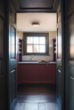 Through a tall archway of a grand, refectory red classic English kitchen is a small scullery room featuring a sink run with Carrara marble worktop, an embedded brass sink, and a brass tap, open counter cupboards, and pantry cupboards. Place upon a tall, magnificent open space with deep olive green walls with ornate gold gilding, and beautifully decorated with a Gaselier light, artisan ceramics, and vintage furniture, creating a splendidly royal and practical use of space.