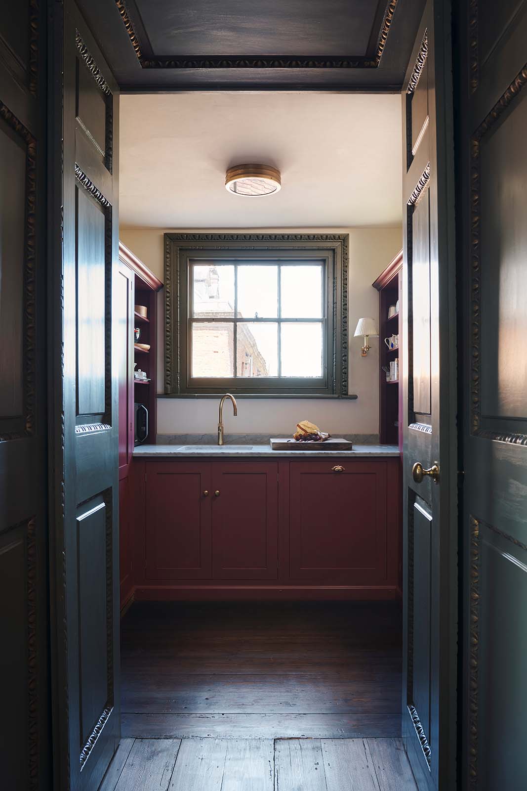 Through a tall archway of a grand, refectory red classic English kitchen is a small scullery room featuring a sink run with Carrara marble worktop, an embedded brass sink, and a brass tap, open counter cupboards, and pantry cupboards. Place upon a tall, magnificent open space with deep olive green walls with ornate gold gilding, and beautifully decorated with a Gaselier light, artisan ceramics, and vintage furniture, creating a splendidly royal and practical use of space.