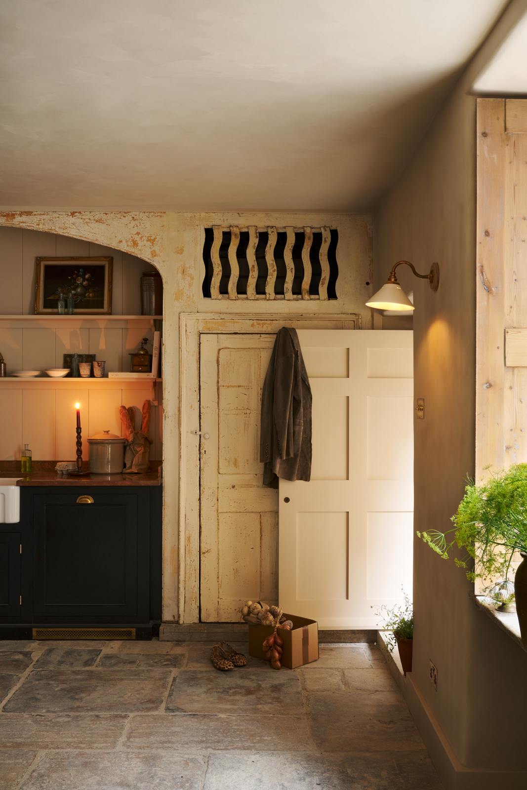 Looking through an open door of a rustic basement kitchen with sandy-coloured walls and ceiling, original Georgian detailing, cream doors and panelling with chipped paintwork and a worn stone floor. On the left is an alcove containing a small run of painted black cupboards with a mottled copper worktop, and a fluted Belfast sink below shelves decorated with vintage bric-a-brac. On the right is an heirloom wall light illuminating.