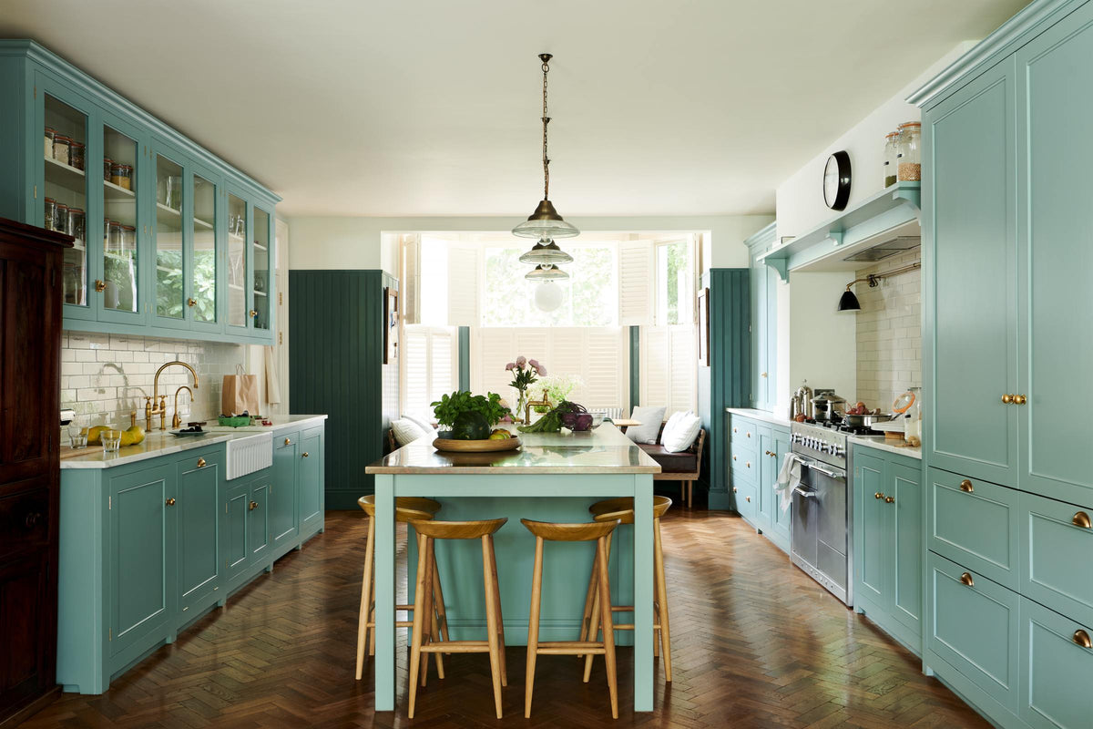 A spacious yet modest Trinity blue classic English kitchen features a Silestone worktop with a sink and cooker run, glazed cupboards, a full-height cabinet with drawers, a kitchen island with a copper sink, aged brass Mayan taps, a copper prep table, and deVOL wooden bum stools, all topped off with a white porcelain splashback. Lovingly decorated with pink peonies in a vase, glass pendant lights, a watermelon, beetroots, live basil, and dried ingredients in glass jars, the room feels humble and warm.