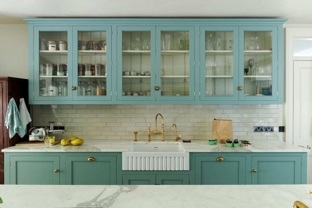 A spacious yet modest Trinity Blue classic English kitchen features a Silestone worktop sink run with a single fluted Belfast sink and aged brass Ionian tap, glazed cupboards, and a kitchen island with a copper sink—all topped off with a white porcelain splashback. Lovingly decorated with a silver juicer, papayas, live basil, berries, dried ingredients in glass jars, the room feels humble, warm, and inviting.