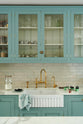 A spacious yet modest Trinity Blue classic English kitchen features a Silestone worktop sink run with a single fluted Belfast sink and aged brass Ionian tap, glazed cupboards, and a kitchen island with a copper sink—all topped off with a white porcelain splashback. Lovingly decorated with a silver juicer, papayas, live basil, berries, dried ingredients in glass jars, the room feels humble, warm, and inviting.