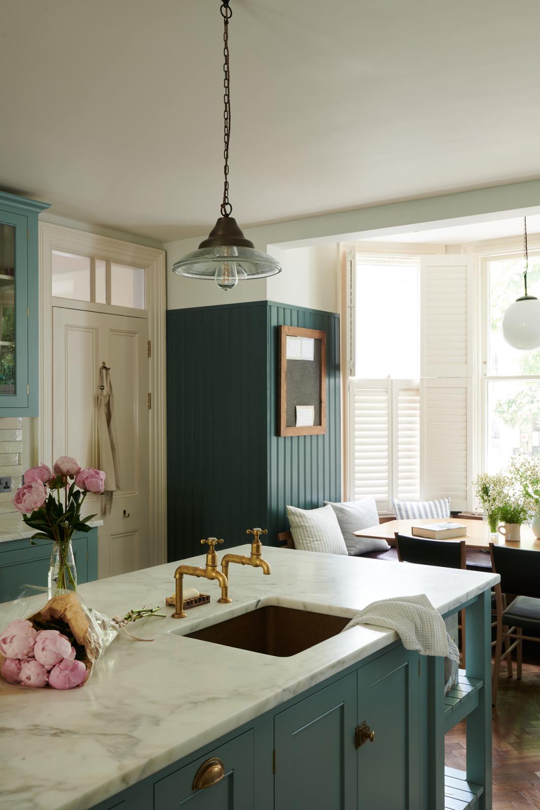 A close, angled view of a spacious yet modest Trinity Blue classic English kitchen features a marble worktop island with a copper sink and aged brass Mayan taps, alongside a breakfast table by the arched window. Lovingly decorated with pink peonies in a vase, glass pendant lights, and a small blackboard and book, the room feels humble, warm, and inviting.