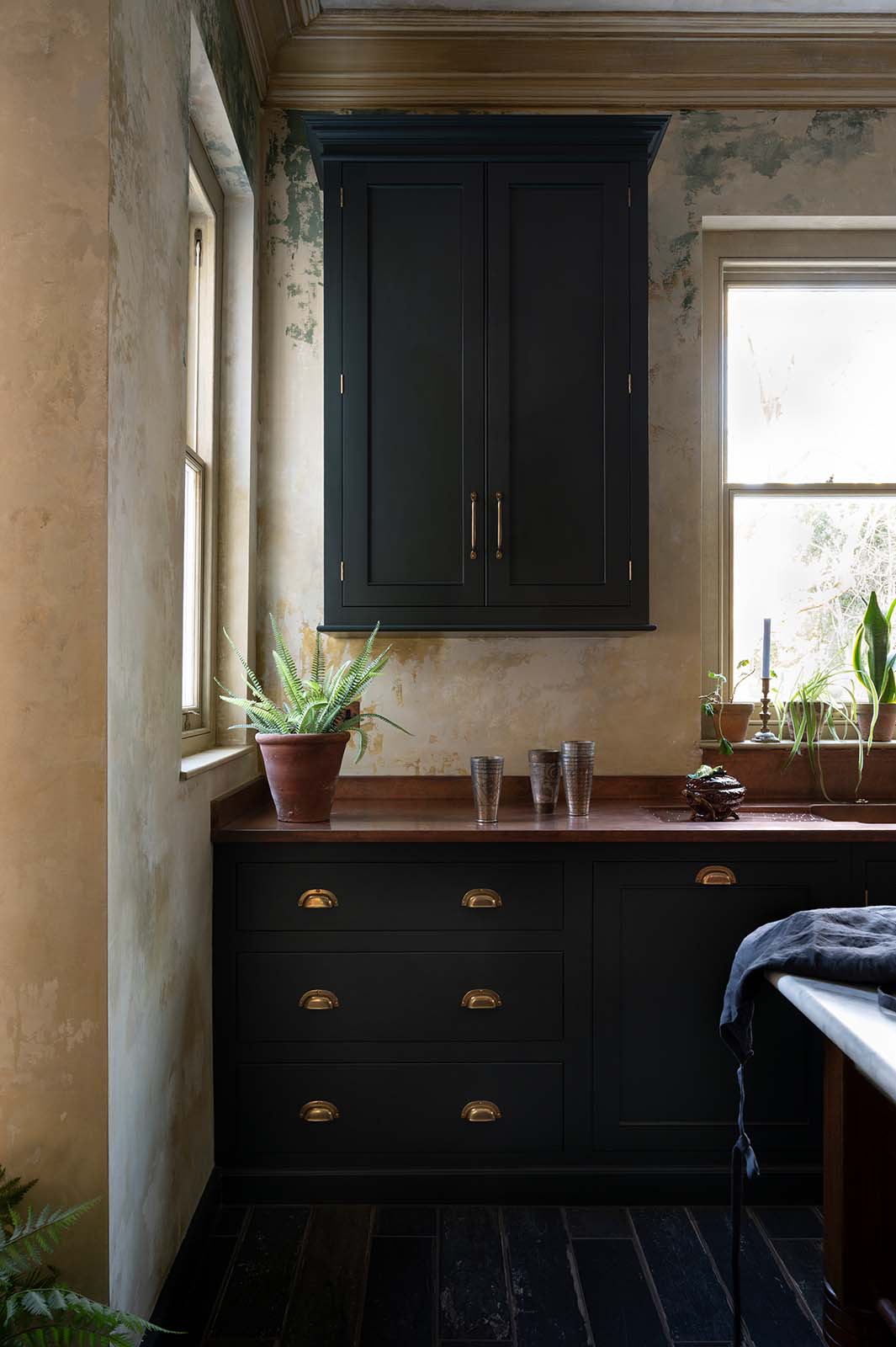 A closer look at an elegant classic kitchen’s sink run reveals an aged copper worktop, wall cupboards, and a marble worktop heirloom dairy table. The pantry-blue wall cupboard takes centre stage, providing functional storage even in the corner of the tall-ceilinged kitchen.