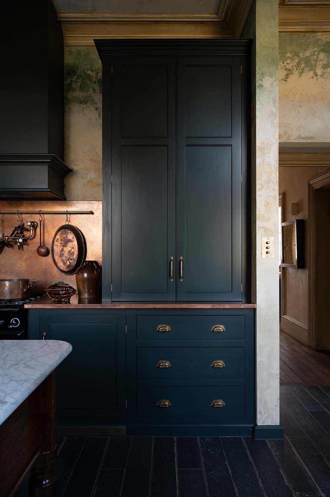 A closer look at an elegant classic kitchen’s cooker run reveals an aged copper worktop, a shaker canopy, counter cabinets, and a marble worktop heirloom dairy table. The pantry-blue counter cabinets stand tall, and beside them is a black gas cooker, copper pans, and fresh chillies hanging from aged brass rails, with a large aged copper splashback behind.