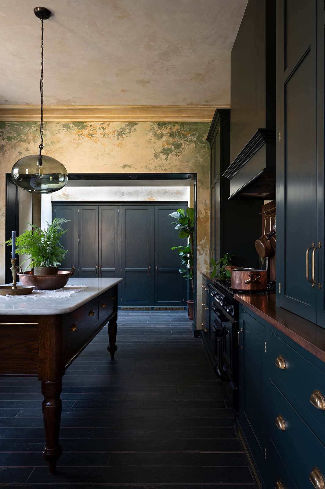An elegant classic kitchen includes a cooker with an aged copper worktop, a shaker canopy, counter cabinets, and a marble worktop heirloom dairy table. The tall-ceilinged room has a rustic wash finish, with green draping down the walls, resembling overgrown ancient ruins. Beyond the archways leading to the hallway is a large classic English fitted larder, perfect for storage or general pantry use.