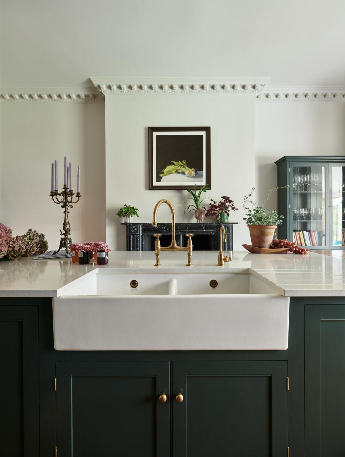 A closer look at the long kitchen island’s double Belfast sink featuring a Silestone worktop and an aged brass Ionian tap in a classic English kitchen design, finished in grand bakehouse green. Adorned with white and blush pink hydrangeas, potted plants, still life paintings, a candelabra, and homemade jams, the room feels homey and welcoming despite its large and decorative appearance.