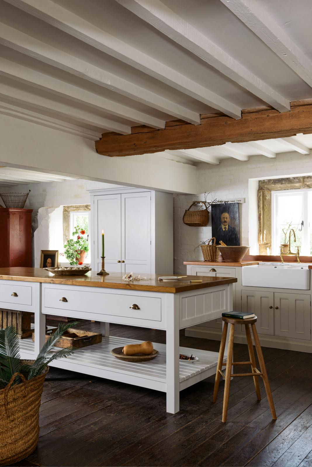 An angled view of a beautiful white linen kitchen featuring a sink run with an aged brass Ionian tap, a freestanding table, a pantry cupboard, and a collection of vintage furniture and items reflecting on the well-reserved historical space with its original plastered walls, wooden beams and oak floors.