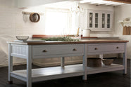An angled view of a beautiful white linen kitchen featuring a sink run with aged brass Ionian taps, a freestanding table, and a collection of vintage furniture and items reflecting on the well-reserved historical space with its original plastered walls, wooden beams and oak floors.